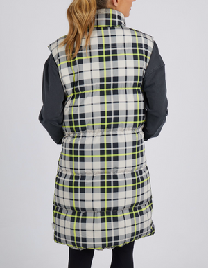 Seen from behind, a person with long hair is wearing a Foxwood Check It Long Line Vest in white, black, and yellow over a dark long-sleeve shirt against a plain light background.