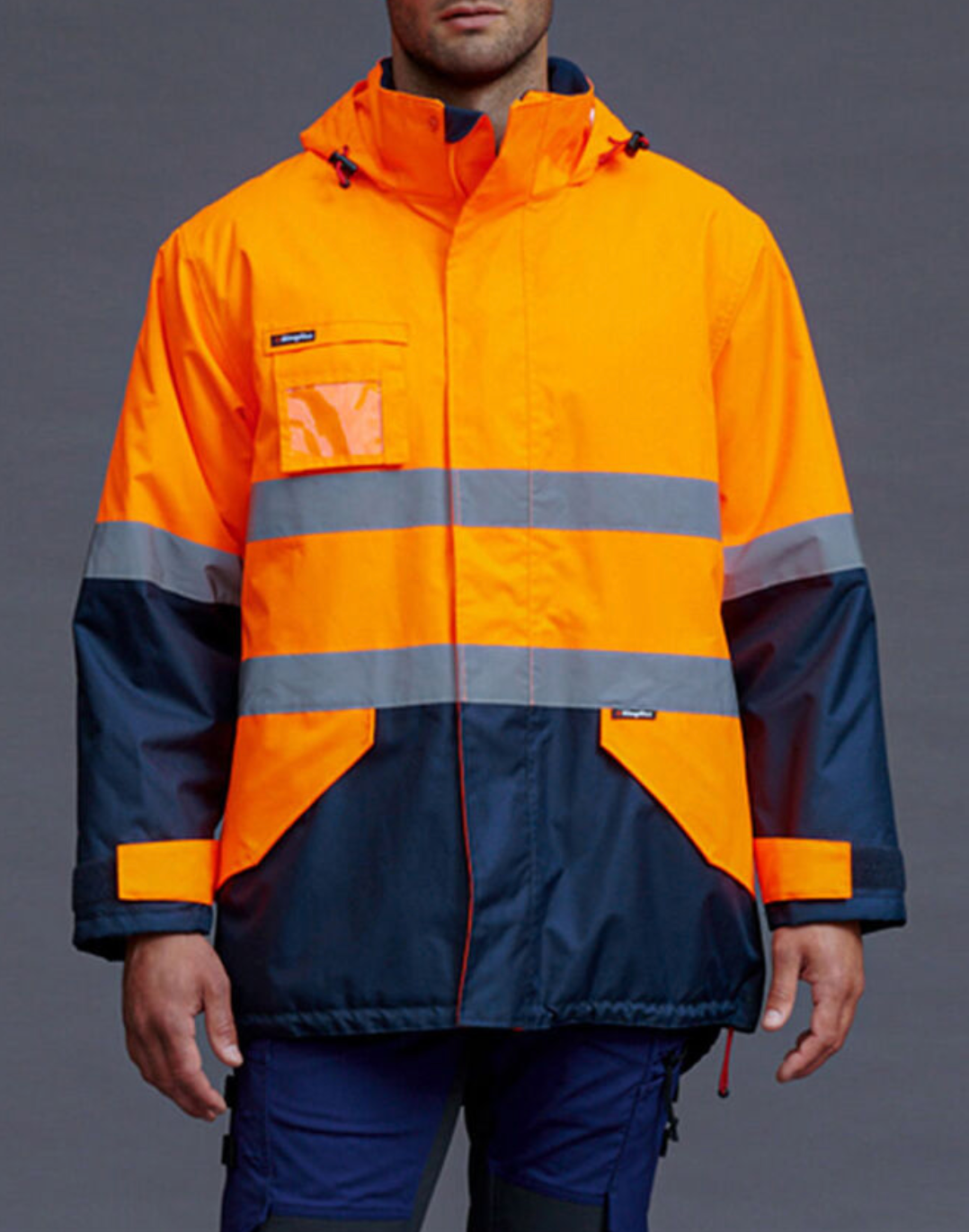 A person wears the King Gee Waterproof Hi Vis Reflective Spray Jacket in bright orange and navy, featuring gray reflective tape and an adjustable hood, standing against a plain gray background.