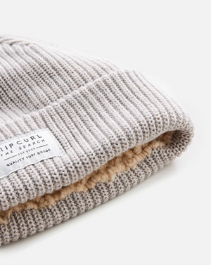 A close-up of the Rip Curl Dusk Fleece Lined Regular Beanie, showcasing its light grey rib knit, soft beige fleece lining, and a white "RIP CURL THE SEARCH" label on the folded edge.