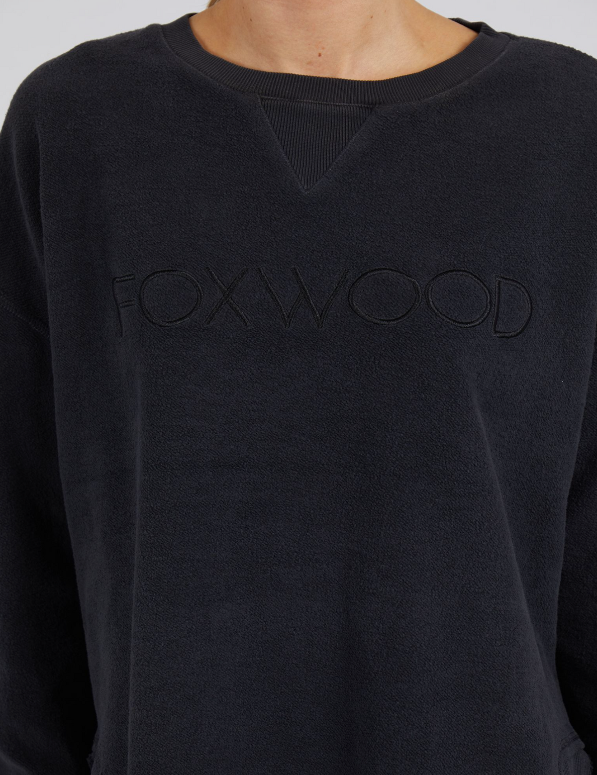 A person is cozy in the Foxwood Cozy Simplified Crew, a dark sweatshirt with an embossed logo on the front, made from teddy fleece for unparalleled softness and comfort.
