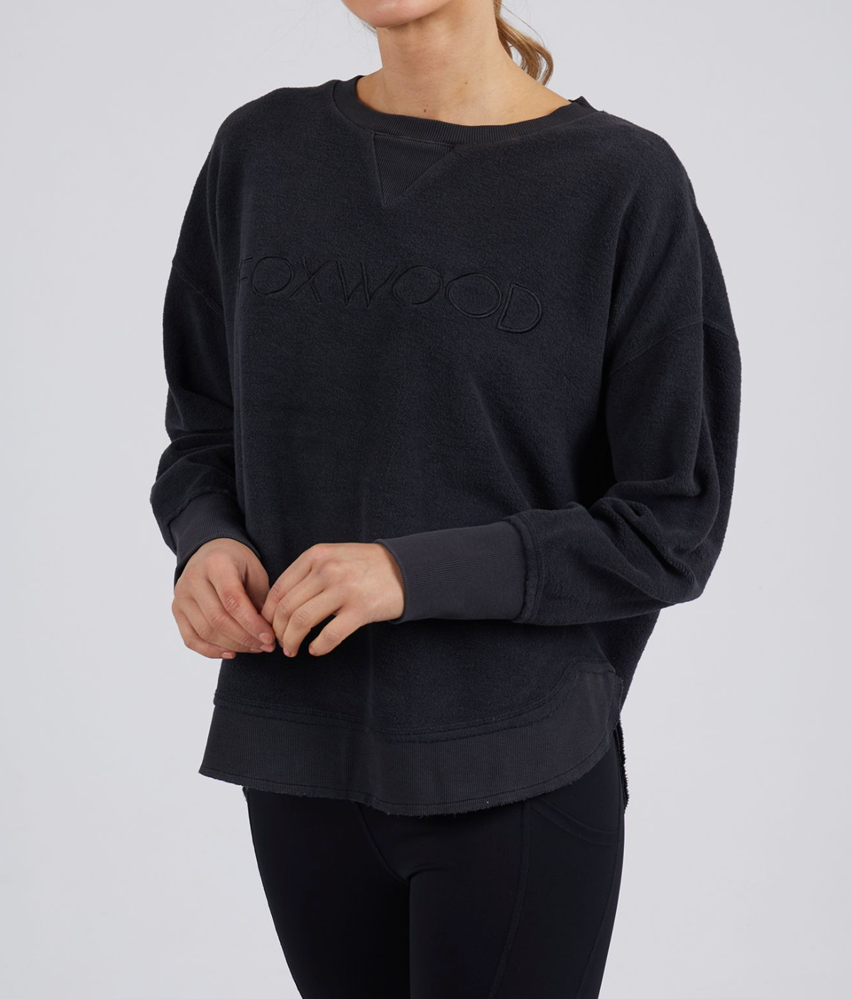 Dressed in a cozy Foxwood Simplified Crew sweatshirt embossed with "OXWOOD" and black leggings, they stand with hands clasped against a plain white background, exuding effortless style and comfort.