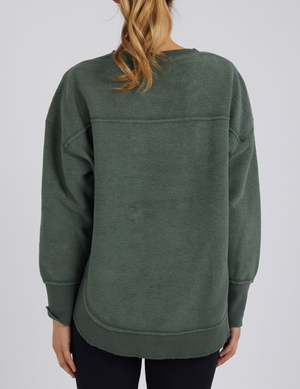 A person with long blonde hair in a ponytail is seen from behind, wearing the oversized Foxwood Cozy Simplified Crew in dark green and black pants, showcasing a relaxed, casual look against a simple light backdrop.