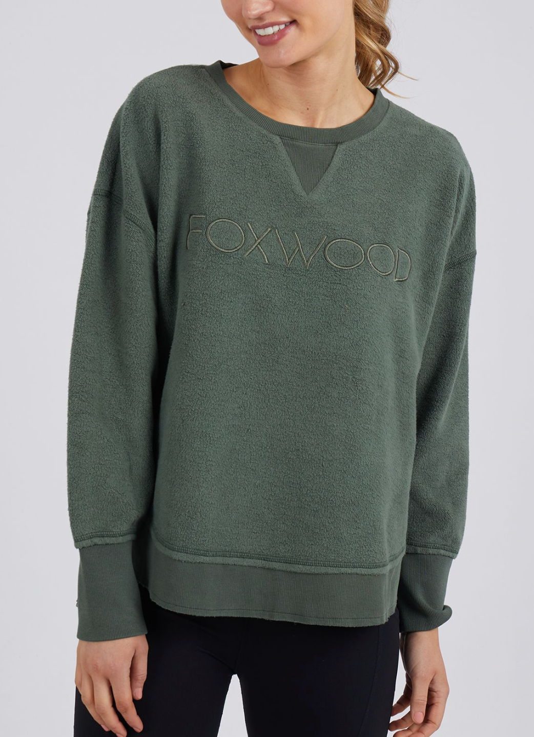 A woman smiles with her head slightly tilted, wearing the Foxwood Cozy Simplified Crew, a casual loose-fit forest green sweatshirt with "FOXWOOD" embroidery on the front, paired with black leggings.