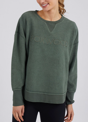 A woman smiles with her head slightly tilted, wearing the Foxwood Cozy Simplified Crew, a casual loose-fit forest green sweatshirt with "FOXWOOD" embroidery on the front, paired with black leggings.