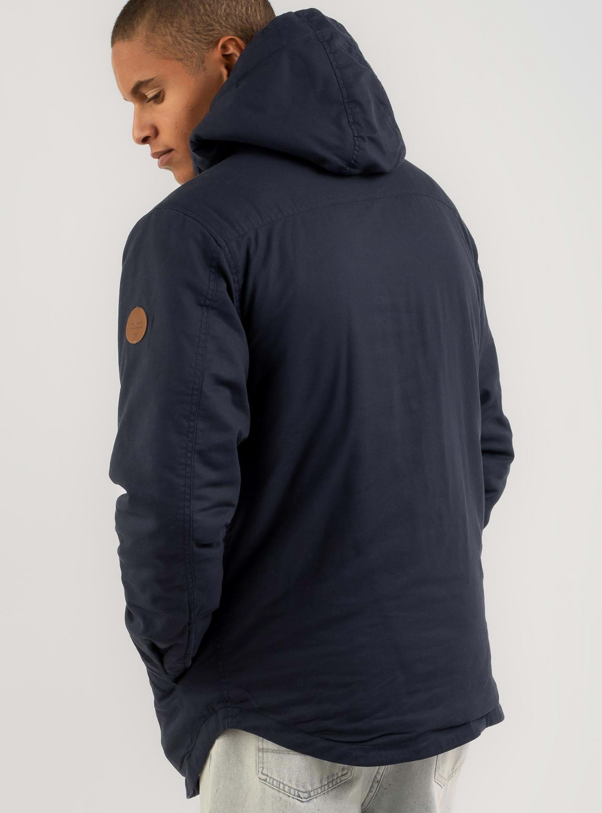 A person in a navy Rip Curl Gibbos Jacket with sherpa lining and a brown circular patch on the upper arm stands facing away from the camera with hands in pockets, wearing light blue jeans.