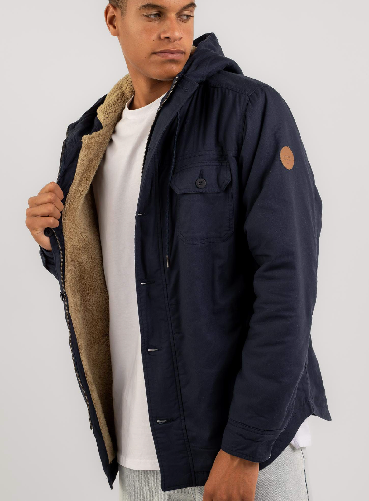 A man in a Rip Curl Gibbos Jacket, dark blue, with tan faux-fur lining and made from a cotton/polyester blend, stands against a plain background. He casually holds the jacket open over his white T-shirt, gazing thoughtfully to the side.