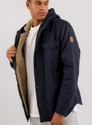 A man in a Rip Curl Gibbos Jacket, dark blue, with tan faux-fur lining and made from a cotton/polyester blend, stands against a plain background. He casually holds the jacket open over his white T-shirt, gazing thoughtfully to the side.