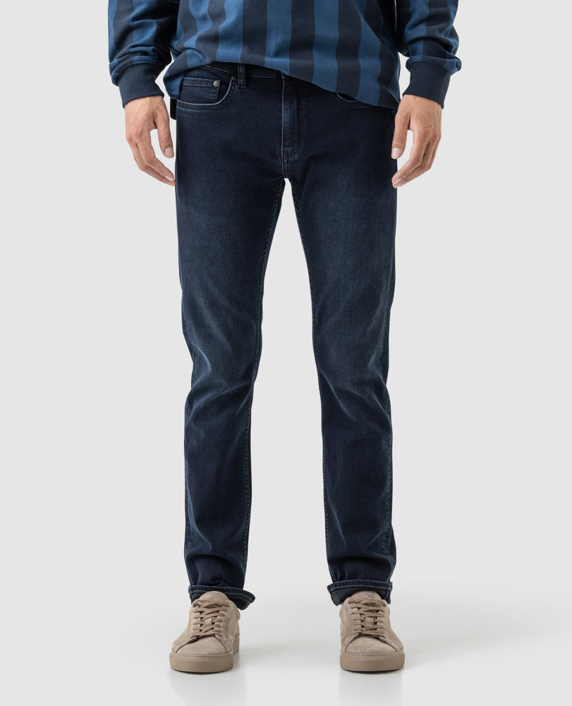 A person wearing Rodd & Gunn Riverstone Slim Jean is standing against a plain background in a blue striped sweater and beige sneakers, with the focus on the lower half of their body.