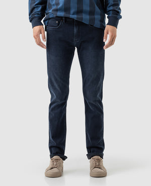 A person wearing Rodd & Gunn Riverstone Slim Jean is standing against a plain background in a blue striped sweater and beige sneakers, with the focus on the lower half of their body.