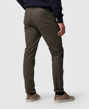 Rear view of a person wearing Rodd & Gunn Straight Fit Jean in dark gray, paired with a dark blue sweater and light brown suede sneakers. The person is standing with their right arm slightly bent and their left hand resting on their thigh against a light gray background.