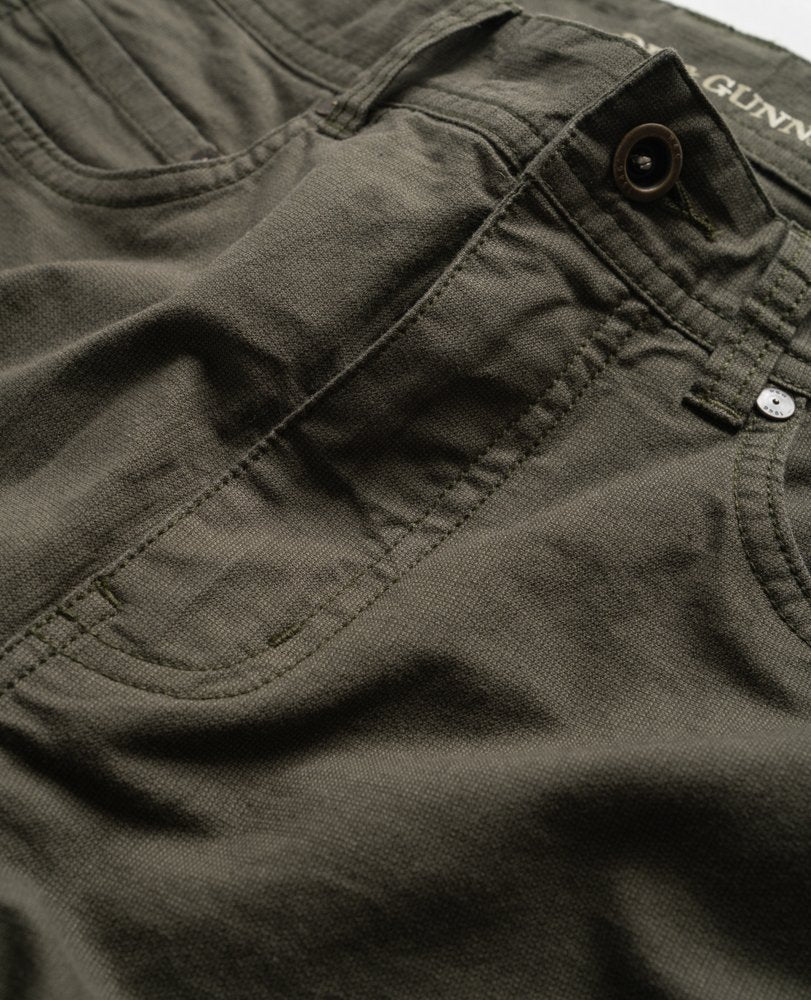 A close-up of dark green Rodd & Gunn Straight Fit Jeans showing the front pocket, waistband, and button. The fabric appears to be a sturdy, heavy material with visible stitching and a buttonhole. The brand name "Gunn" can be partially seen on the inside waistband, complemented by antique brass hardware.