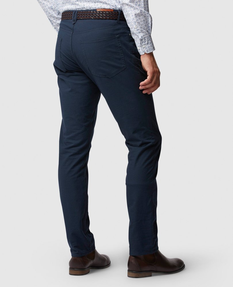 A person is standing and facing away, wearing a long-sleeve patterned shirt, a woven leather belt, comfortable Rodd & Gunn Straight Fit Jeans with a mid-rise waist, and brown dress shoes. The background is plain white.