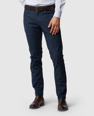 A person dressed in a white patterned shirt is modeling the Rodd & Gunn Straight Fit Jean in dark blue, which features a mid-rise waist and is made of stretch fabric, paired with a brown braided belt. The outfit is completed with brown leather boots. The background is plain white, and the person's face is not visible in the image.