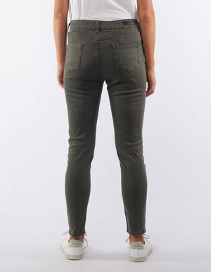 Back view of a person wearing Foxwood City Jean in olive green and a white t-shirt. The jeans feature two back pockets and are slightly cropped above the ankles. The individual is also sporting white Veja sneakers with the brand's logo visible on the heel. The background is white.