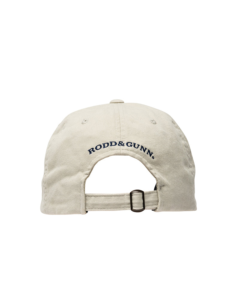 The image shows the back view of the Rodd & Gunn Signature Cap, a beige cotton twill baseball cap with an adjustable strap. The brand name "RODD & GUNN" is embroidered above the strap in navy blue letters, marking it as a standout piece from the stylish brand.