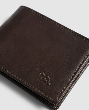 A close-up image of a dark brown Rodd & Gunn Victoria Flats Road Wallet with visible stitching. The genuine buffalo leather wallet features an embossed design of a dog on its lower right corner. The background is light grey.