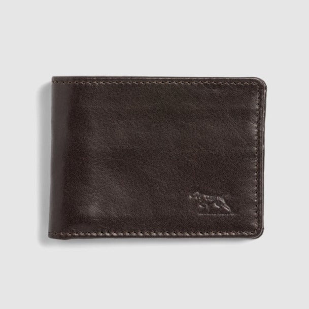 The Rodd & Gunn Victoria Flats Road Wallet, in a closed configuration, boasts a dark brown hue and is meticulously crafted from genuine buffalo leather with prominent stitching along the edges. An embossed animal logo is subtly positioned on the lower right corner. The background is light gray.