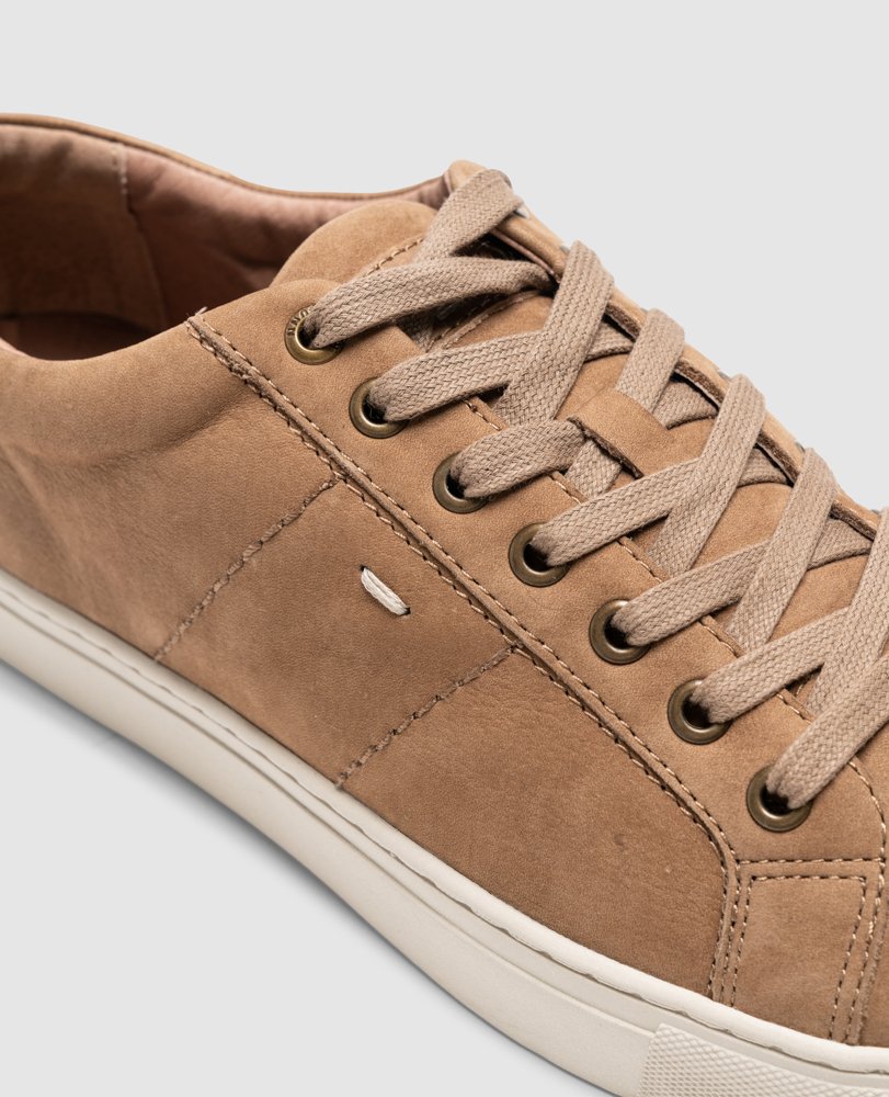Close-up of the Rodd & Gunn Endeavour Spirit Sneaker in tan suede, crafted from nubuck leather with beige laces and metal eyelets. Detailed stitching and a small embossed logo complement the white sole, highlighting its clean, minimalist design against a light gray background.