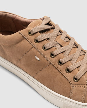 Close-up of the Rodd & Gunn Endeavour Spirit Sneaker in tan suede, crafted from nubuck leather with beige laces and metal eyelets. Detailed stitching and a small embossed logo complement the white sole, highlighting its clean, minimalist design against a light gray background.