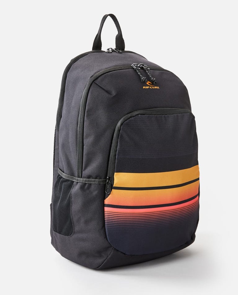 The Rip Curl Ozone 30L School Eco Backpack, made from recycled plastic, sports a black design with vibrant orange, pink, and yellow gradient stripes on the front pocket. It has two zippered compartments, a laptop compartment, top handle, and side mesh pockets for essentials.