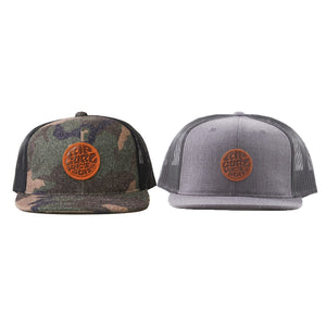 Two Rip Curl Premium Wetty Trucker caps are displayed side by side. The cap on the left features a camouflage pattern, while the one on the right is solid grey with a mesh back. Both caps have adjustable straps and are adorned with a round leather patch with a logo at the center front.