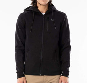 A person with long hair is wearing a black Rip Curl Anti Series Departed Zip Thru hoodie with drawstrings and beige pants. The hoodie, perfect for winter clothing, features a subtle logo on the left chest pocket and another one on the left sleeve. Its Durable Water Resistant (DWR) coating adds extra protection. The background is plain white.