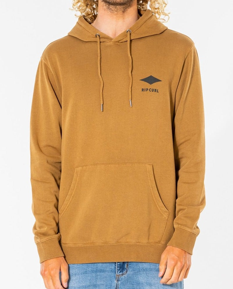 A person wears a brown Rip Curl Quality Products Hood, made from a cozy cotton/polyester blend, featuring the logo and text on the chest, with a front pocket and drawstrings. They're paired with light blue jeans against a plain white background.