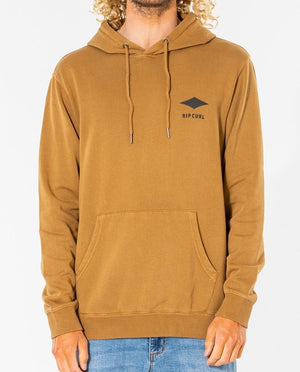 A person wears a brown Rip Curl Quality Products Hood, made from a cozy cotton/polyester blend, featuring the logo and text on the chest, with a front pocket and drawstrings. They're paired with light blue jeans against a plain white background.