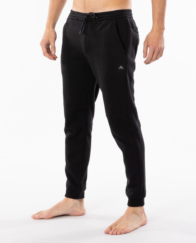 A person stands barefoot wearing the Rip Curl Departed Anti Series Trackpant in black. These jogger pants feature an elastic waistband with a drawstring, side pockets, and a small logo on the left thigh. Perfect for winter clothing needs, they offer comfort against a plain white background.