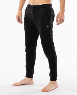 A person stands barefoot wearing the Rip Curl Departed Anti Series Trackpant in black. These jogger pants feature an elastic waistband with a drawstring, side pockets, and a small logo on the left thigh. Perfect for winter clothing needs, they offer comfort against a plain white background.