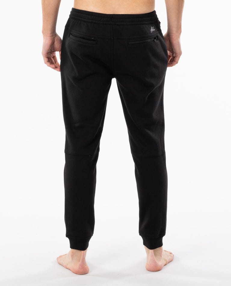 Rear view of a person from the waist down, wearing Rip Curl Departed Anti Series Trackpant in black. The person is standing barefoot on a plain white background. These joggers, ideal for winter wear, feature visible back pockets with zippers and are crafted from durable water-resistant material.