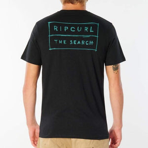 A person wearing a black Rip Curl 50/50 Scratch Tee, featuring "THE SEARCH" printed in teal on the back and made from 100% cotton for comfort, is paired with khaki pants. The person has light hair.