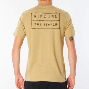 Back view of a person wearing a light brown Rip Curl 50/50 Scratch Tee, made from 100% cotton, featuring the text "RIP CURL THE SEARCH" printed in a rectangular box on the back. The comfortable tee reveals a tattoo of an eye on their left forearm.