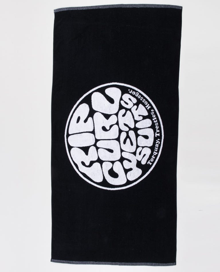 Introducing the Rip Curl Wetty Icon Towel: a black cotton jacquard towel featuring a white circular logo at its center with stylized text inside the circle that reads "Rip Curl, Wetty" and smaller text along the edge of the circle that reads "Torquay, Hossegor." The towel is finished with gray trim along its edges.