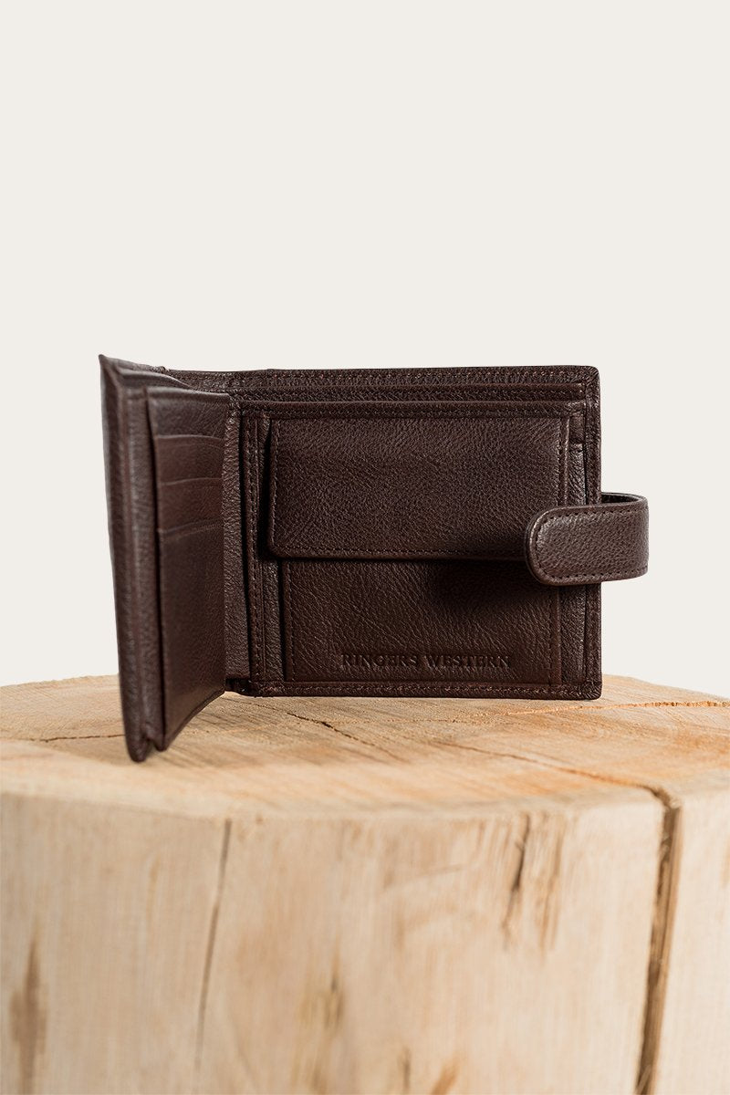 The Ringers Western Cypress Wallet, made of genuine leather, is elegantly displayed on a wooden block against a plain backdrop. This refined wallet includes card slots and a small flap compartment secured with a snap closure.
