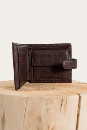 The Ringers Western Cypress Wallet, made of genuine leather, is elegantly displayed on a wooden block against a plain backdrop. This refined wallet includes card slots and a small flap compartment secured with a snap closure.
