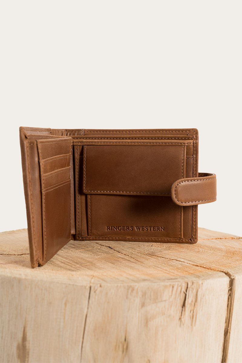 The Ringers Western Cypress Wallet, made from genuine brown leather, features a clasp and iconic logo. Inside, find multiple card slots and a coin pocket. Elegantly displayed on a wooden surface with a light background.