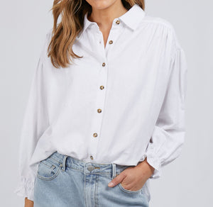A person with long hair is wearing a white, long-sleeved Foxwood Mia Shirt crafted from a viscose linen blend, paired with light blue jeans. The shirt features brown buttons and slightly puffed sleeves with elastic cuffs. The background is plain white.