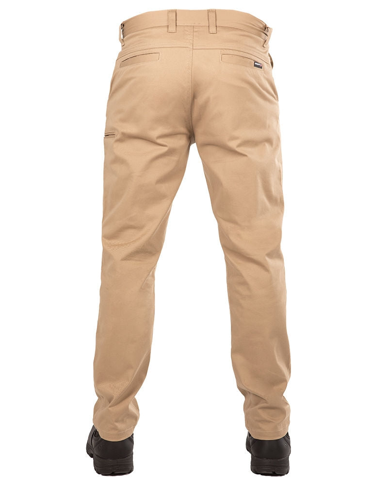 A person wearing UNIT Ignition Stretch Pants in tan and black boots is standing with their back to the camera. The durable work pants, made from stretch fabric, feature pockets, belt loops, a visible brand label near the waist, and a small utility pocket on the left pant leg.
