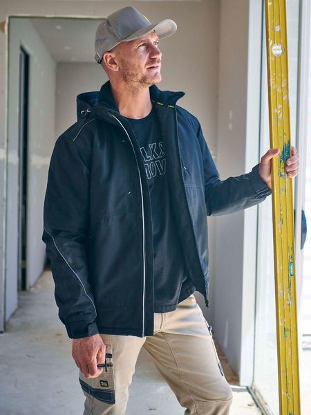 A man in a gray cap, beige pants, and a Bisley Heavy Duty Dobby Jacket with an adjustable hood stands in a partially constructed room. He holds and looks up at a yellow level tool, surrounded by unfinished walls and an open doorway in the background.