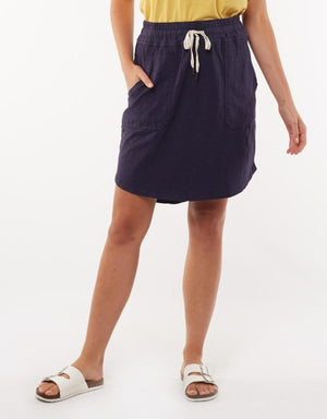 A person is standing against a plain background, dressed in a yellow top tucked into a Foxwood Utility Skirt. This comfortable navy skirt, made from 100% cotton, features an elastic waistband and drawstring. The person's hands are in the skirt's pockets, and they are also wearing white sandals.