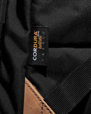 Close-up of a black fabric label on the FXD WBP-3 Work Backpack, showcasing "CORDURA Ballistic fabric" in orange and white. The label is sewn onto black material with a partially visible brown leather base.