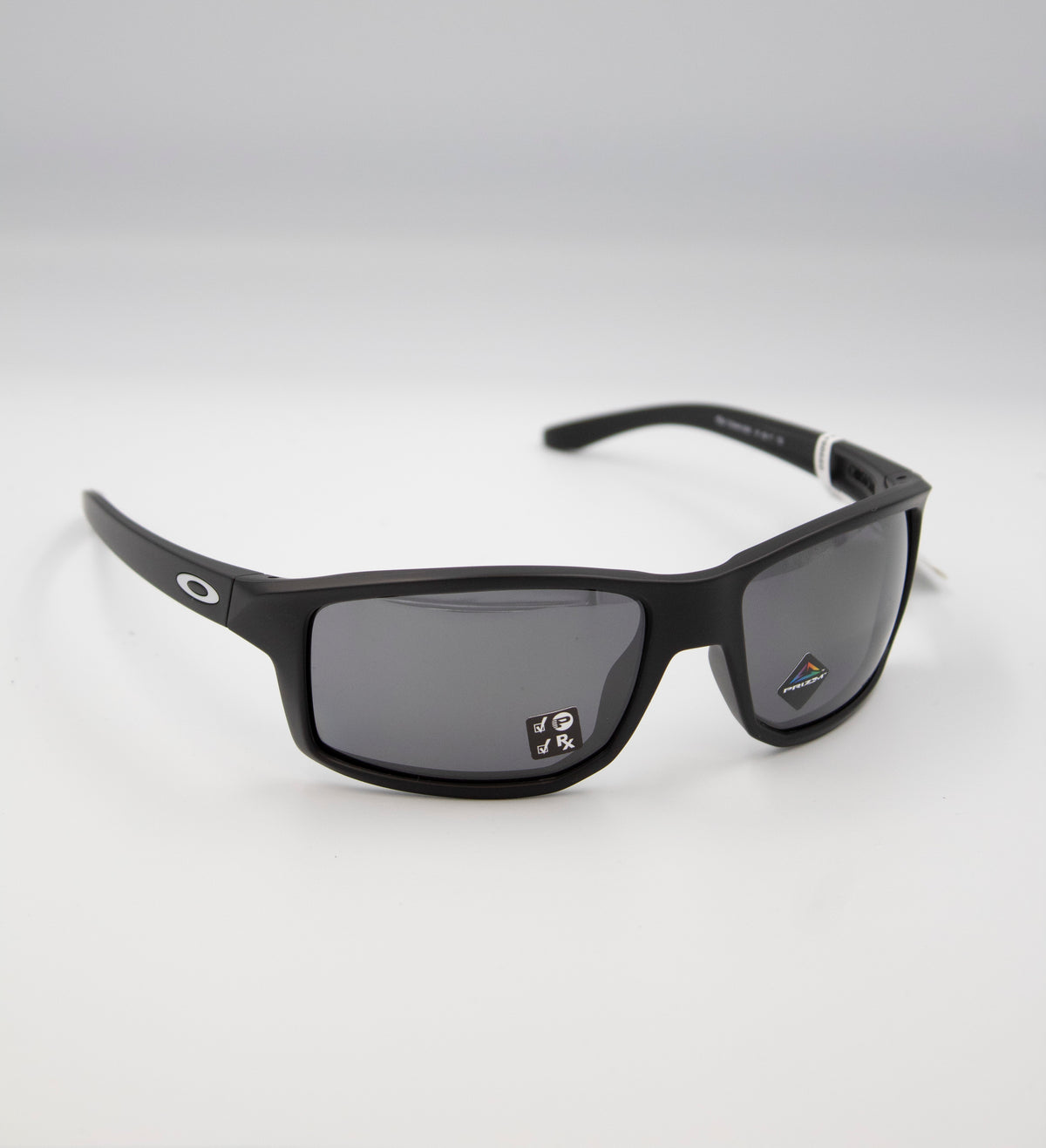 A pair of sleek Oakley Gibston Polarized sunglasses in matte black with gray lenses, featuring the iconic Oakley logo on the arms. Positioned on a white surface with a subtle shadow, they have a small sticker on one lens.