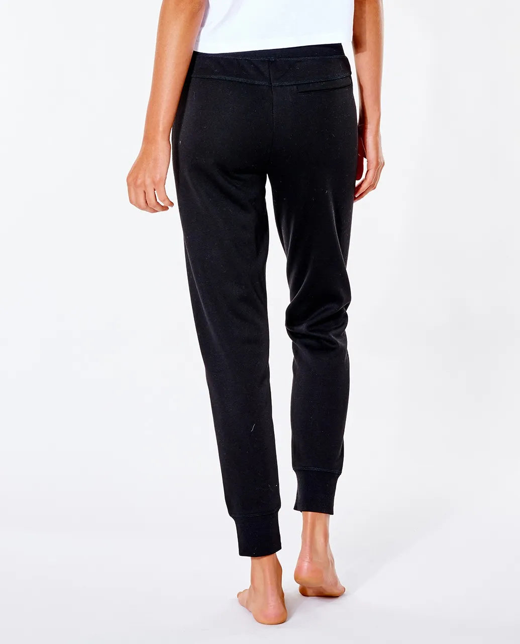 A person wearing a white shirt and black Rip Curl Anti Series Flux II Trackpant, shown from the back. The person's feet are bare, and the background is plain white. The trackpants have a fitted design with ribbed cuffs at the ankles, making them ideal winter clothing featuring durable water-resistant fabric.