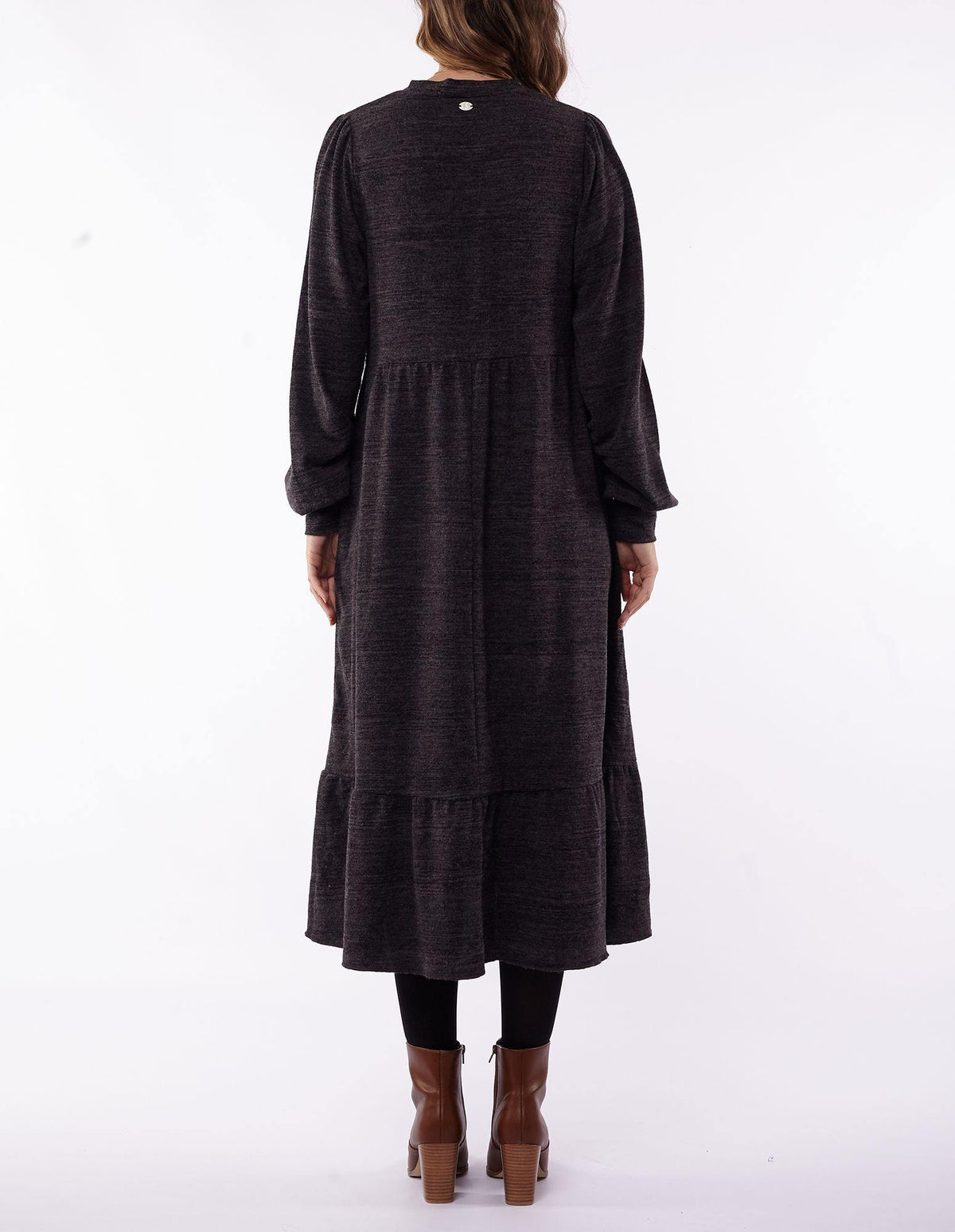 Back view of a person wearing the Foxwood Kensington Dress, a long-sleeved, dark gray midi-length dress with a tiered hem, paired with black leggings. The stylish dress is complemented by brown ankle boots and perfectly matches the person's long hair. The background is white.