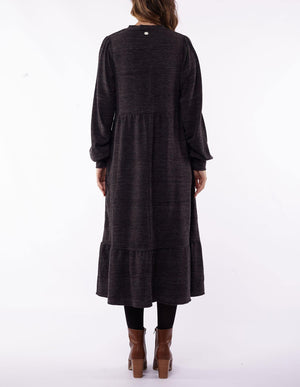 Back view of a person wearing the Foxwood Kensington Dress, a long-sleeved, dark gray midi-length dress with a tiered hem, paired with black leggings. The stylish dress is complemented by brown ankle boots and perfectly matches the person's long hair. The background is white.