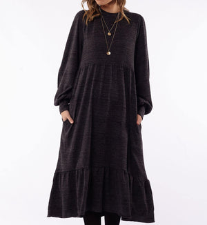 A person wearing the Foxwood Kensington Dress, a long, stylish piece in dark gray with long sleeves and a ruffled hem. The comfortable dress features pockets and is accessorized with a long necklace adorned with two circular pendants. The background is plain white, and the person's head is not visible in the image.