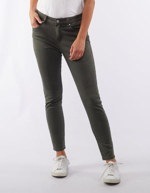 A person standing against a plain white background, wearing a white t-shirt, Foxwood City Jean in olive green, and white sneakers. The person has one hand in a pocket and the other rests casually by their side.