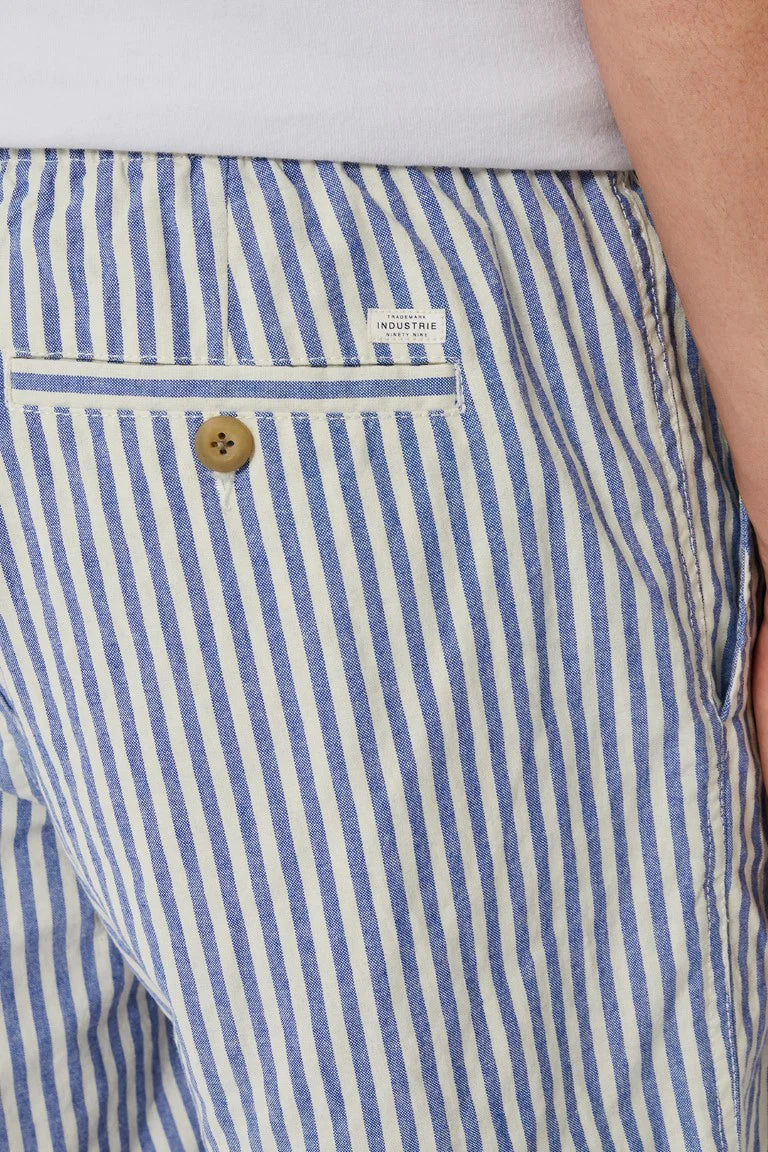 A close-up shows someone wearing Industrie The Laguna Short, featuring stylish blue and white stripes and an elastic waist. The shorts have a back pocket with a button and a white label, while the person's hand partially appears on the right, enhancing the casual chic vibe.