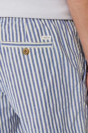 A close-up shows someone wearing Industrie The Laguna Short, featuring stylish blue and white stripes and an elastic waist. The shorts have a back pocket with a button and a white label, while the person's hand partially appears on the right, enhancing the casual chic vibe.